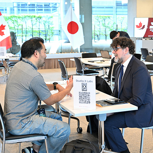 Deux hommes assis à une table mène des discussions d'affaires dans une grande salle avec les drapeaux canadien et japonais en arrière-plan.