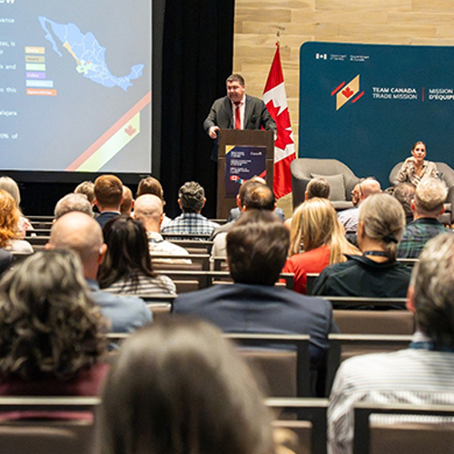 A man at a podium speaks to an audience while two panelists sit in armchairs in front of a banner