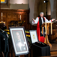 Bishop Shane Parker, wearing his ecclesiastical robes, stands at the lectern. At the forefront, the official Canadian portrait of The Duke. Next to the portrait is the Duke’s cypher and Canadian honours. A uniformed member of the military with a black armband stands next to the portrait.