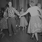 Black and white image of Prince Philip, wearing a checkered shirt and a neck scarf, square dancing at Rideau Hall with other dancers.