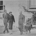 Black and white image of Her Majesty Queen Elizabeth II and The Duke of Edinburgh walking outdoors with Governor General Vincent Massey. Her Majesty and The Duke are looking at the Governor General’s dog Duff, who is walking alongside Mr. Massey and carrying the Queen’s purse.