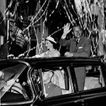 Black and white image of The Duke of Edinburgh and Her Majesty Queen Elizabeth II standing in the back of a convertible car and waving at the crowd. The car is being chauffeur-driven down a busy street. A large amount of ticker tape can be seen falling from above and onto the Royal couple.