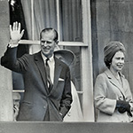 Photo en noir et blanc du duc d’Édimbourg saluant la foule debout à partir d’un balcon, en compagnie de Sa Majesté la reine Elizabeth II.
