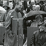 The Duke of Edinburgh standing outside, in front of a crowd, with Lester B. Pearson sitting next to him and laughing.