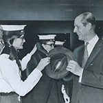 Black and white image of a young girl, wearing the Venturer Scout uniform, exchanging a smile with The Duke of Edinburgh while she is handing him his hat. Other Venturer Scouts are standing in the background.