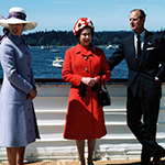 Her Majesty Queen Elizabeth II, Princess Anne and The Duke of Edinburgh are standing together relaxing on the deck of a ship. Water, smaller boats and the coastline can be seen in the background.