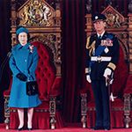 Sa Majesté la reine Elizabeth II et le duc d’Édimbourg debout devant les trônes du Sénat. La Reine porte un manteau et un chapeau bleus, alors que le duc porte un uniforme militaire.