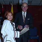 The Duke of Edinburgh standing next to a young girl in traditional dress holding her award. Both are facing forward and smiling.