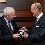Le duc d’Édimbourg sourit alors qu’il reçoit l’insigne de Compagnon de l’Ordre du Canada des mains du gouverneur général David Johnston. Les deux hommes sont debout et portent des costumes d’affaires noirs.