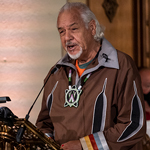 A man wearing a jacket with Indigenous symbols on it speaks at a lectern.