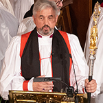 A man, wearing traditional vestments and carrying a crozier, speaks at a lectern.