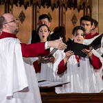 Des choristes disposés en 3 rangées de bancs d’église chantent sous la direction d’un chef de chœur.