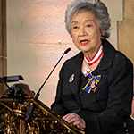 A woman wearing a jacket with decorations speaks at a lectern.