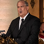 A man wearing a dark suit speaks at a lectern.
