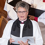 A woman wearing traditional vestments is officiating, observed by choir members sitting in pews behind her.