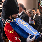 Un homme en tenue militaire et portant un bonnet en peau d’ours noir transporte un drapeau bleu royal plié sur lequel figure la lettre E, l’initiale de la Reine.
