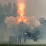 People in military attire stand in a field and observe the resulting flames of a cannon blast.