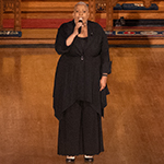 Une femme chante dans le chœur d'une église, observée des choristes et des membres de l'auditoire debout dans des rangs de bancs d'église.