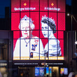 A building at nighttime displays 2 portraits of The Queen, one elderly and one young, on digital screens.