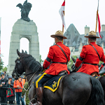 Des gendarmes en tunique rouge défilent à cheval lors d'une parade, observés par une foule le long d'une voie publique passant par un monument.