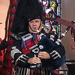 A man, wearing military attire and a bearskin hat, plays the pipe, observed by audience members standing in pews.