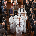 Vue aérienne d’individus en vêtements sacerdotaux défilant dans la nef d’une église, observés de membres de l’auditoire debout dans des rangs de bancs d’église.
