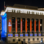 Red and blue projections including a picture of The Queen are projected on a waterfront building at nighttime.