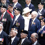 Queen Elizabeth II is seen at a distance, wearing white, and walking among a group of veterans wearing blue uniforms.
