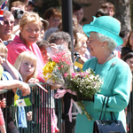 A woman in the crowd taking a picture of Queen Elizabeth II holding flowers.