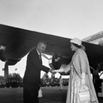 Black and white image of the Queen shaking hands with Prime Minister John Diefenbaker. The Duke of Edinburgh is standing behind her.