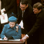 Queen Elizabeth II signing a document surrounded by dignitaries.