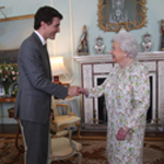 The Queen and Prime Minister Justin Trudeau are standing in front of a fireplace, in a parlor, smiling and shaking hands.