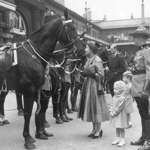 Black and white image of the Queen, along with her two children and an RCMP officer, looking up at a black horse. The Duke of Edinburgh follows behind.