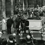 Black and white image of the Queen, greeted at the entrance of a building. The car she exited from is surrounded by a crowd of people.