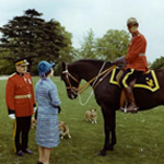Queen Elizabeth II and an RCMP officer are looking up at a horse and its RCMP rider. Two Corgi dogs can be seen in the background.