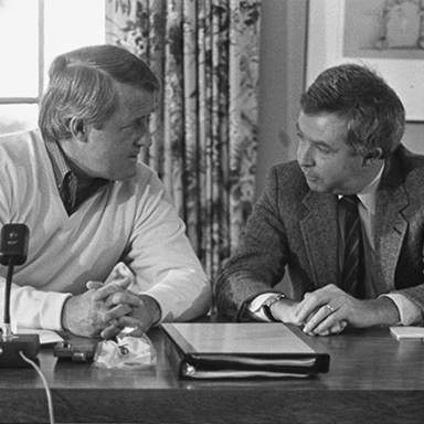 Brian Mulroney and Joe Clark sit side by side at a table while having a discussion.