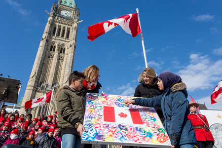 Minister Melanie Joly standing with 3 people, looking at a painting of a Canadian flag, in front of the Parliament Hill Peace tower. 