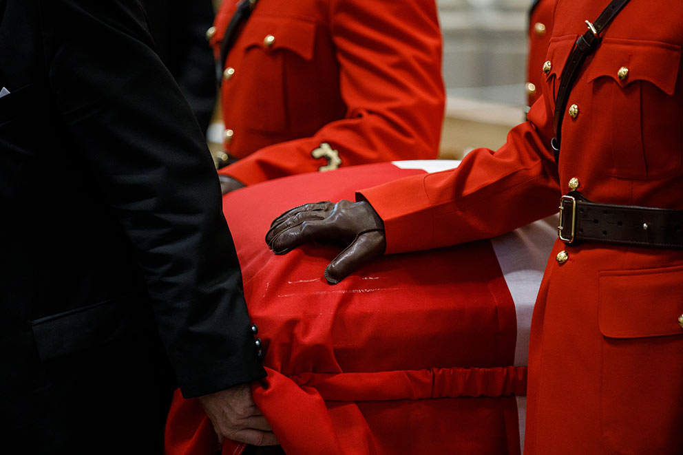 Un porteur en tenue militaire rouge pose une main gantée sur un cercueil.