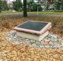 Une plaque de grande taille est fixée sur un socle en pierre au-dessus de galets pavés. Elle est entourée de feuilles d'automne tombées.