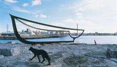 Photo of a bronze sculpture of a boat outline and a bronze sculpture of a dog-wolf perched on a rock by the water.