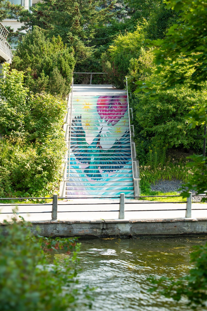 Mural on a staircase set in greenery, depicting a bird with outstretched wings rising toward a pink heart and yellow stars in the sky