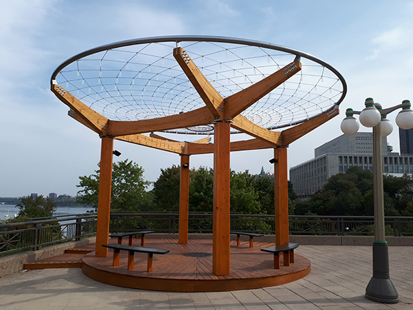 Large ring supported by four wooden pillars. The ring is crisscrossed by metal wires adorned with pieces of glass.