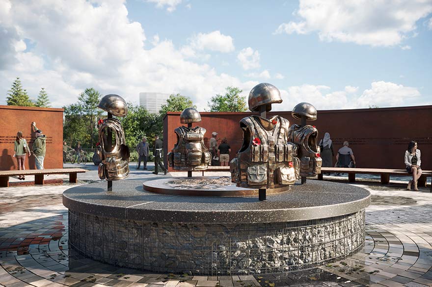 Four flak jackets and helmets are displayed on crosses and arranged in a circle. They sit on top of a raised round stone shape. In the background people are sitting on benches or reading inscriptions on a rust coloured wall. We see green trees and buildings in the distance.