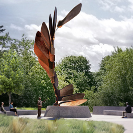 Two people walk along a curved path towards a tall bronze sculpture. The spiraling sculpture is composed of sugar maple seeds swirling in the wind or rooting into the ground.  Lush green trees are in the background.