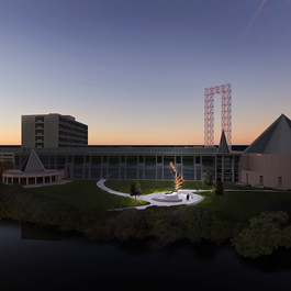 It is sunset. The sky is dark blue with an orange band of light on the horizon. A panoramic view of the site shows an illuminated tall bronze sculpture in front of the Diefenbaker Building. A portion of the river is visible in the foreground.