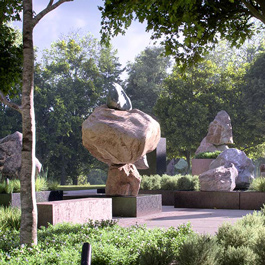 A collection of square plinths topped by boulders sits within a curved plaza. The plaza is surrounded by lawn and bordered on two sides by trees with autumn leaf colours.  
