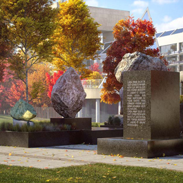 A large black granite block with inscriptions sits in front of boulders of different shapes and sizes. The Diefenbaker Building and trees with autumn leaf colours are visible in the background. 