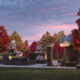 It is sunset; the sky is pink and purple coloured. An ensemble of boulders, blocks and plinths is illuminated. Trees with autumn leaf colours are shown in the background.