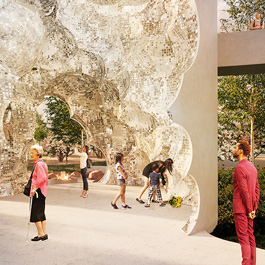 Plusieurs personnes marchent à l'intérieur d'un grand cylindre blanc qui contient une sculpture en morceaux de miroir rappelant un nuage orageux. On voit un aménagement paysager en arrière-plan et des personnes sur une plate-forme surélevée dans le coin supérieur droit.