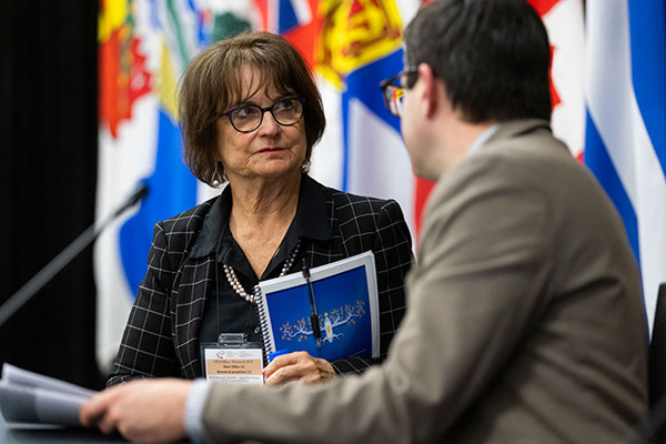 Deborah Lyons, en conversation avec un homme de dos, tient un exemplaire du guide. Derrière eux se trouvent les drapeaux des provinces et territoires canadiens.